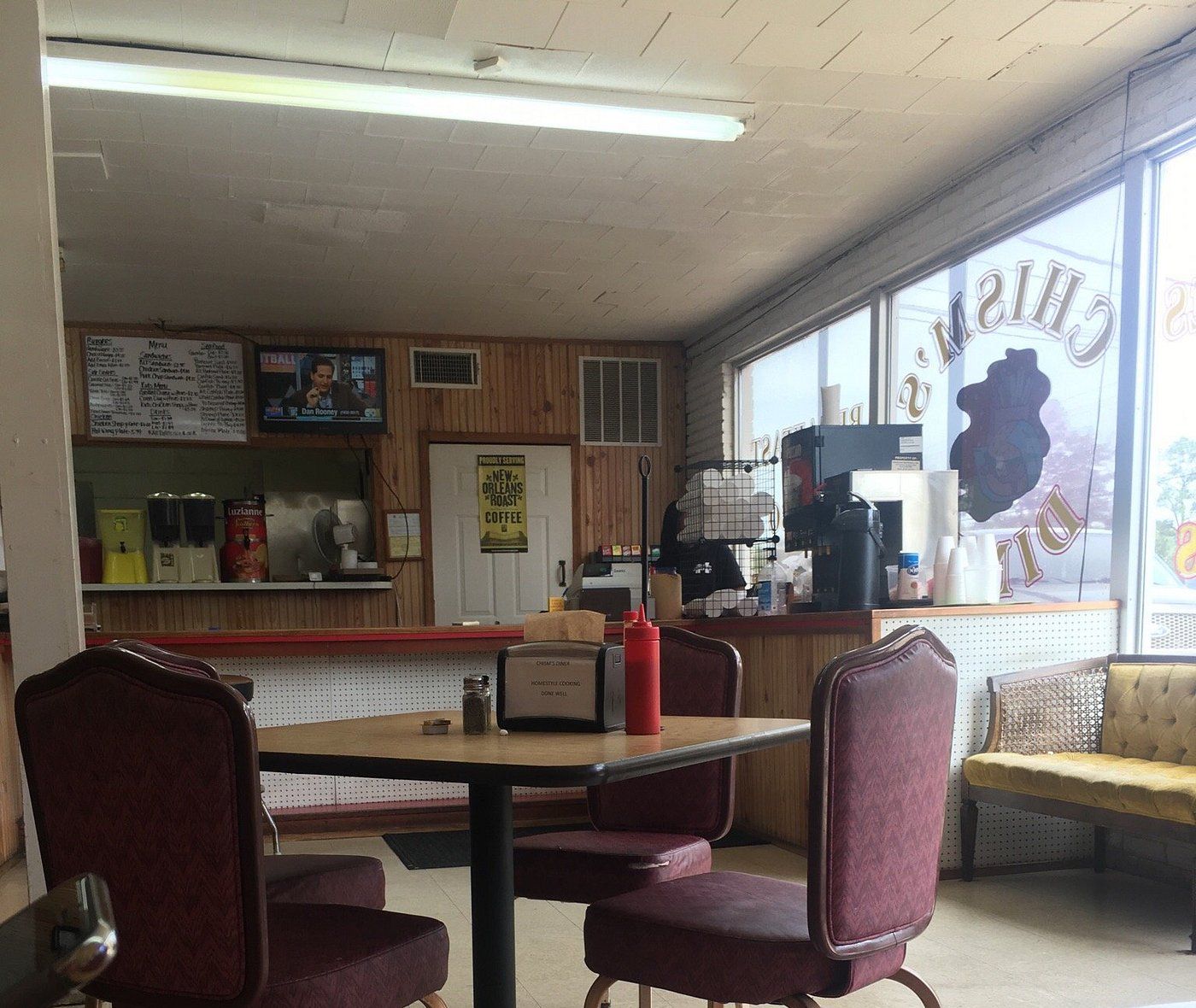 Dining room with burgundy chairs, a square table with a napkin holder, the red-trimmed counter and the Chism's Diner lettering seen backwards on the front window