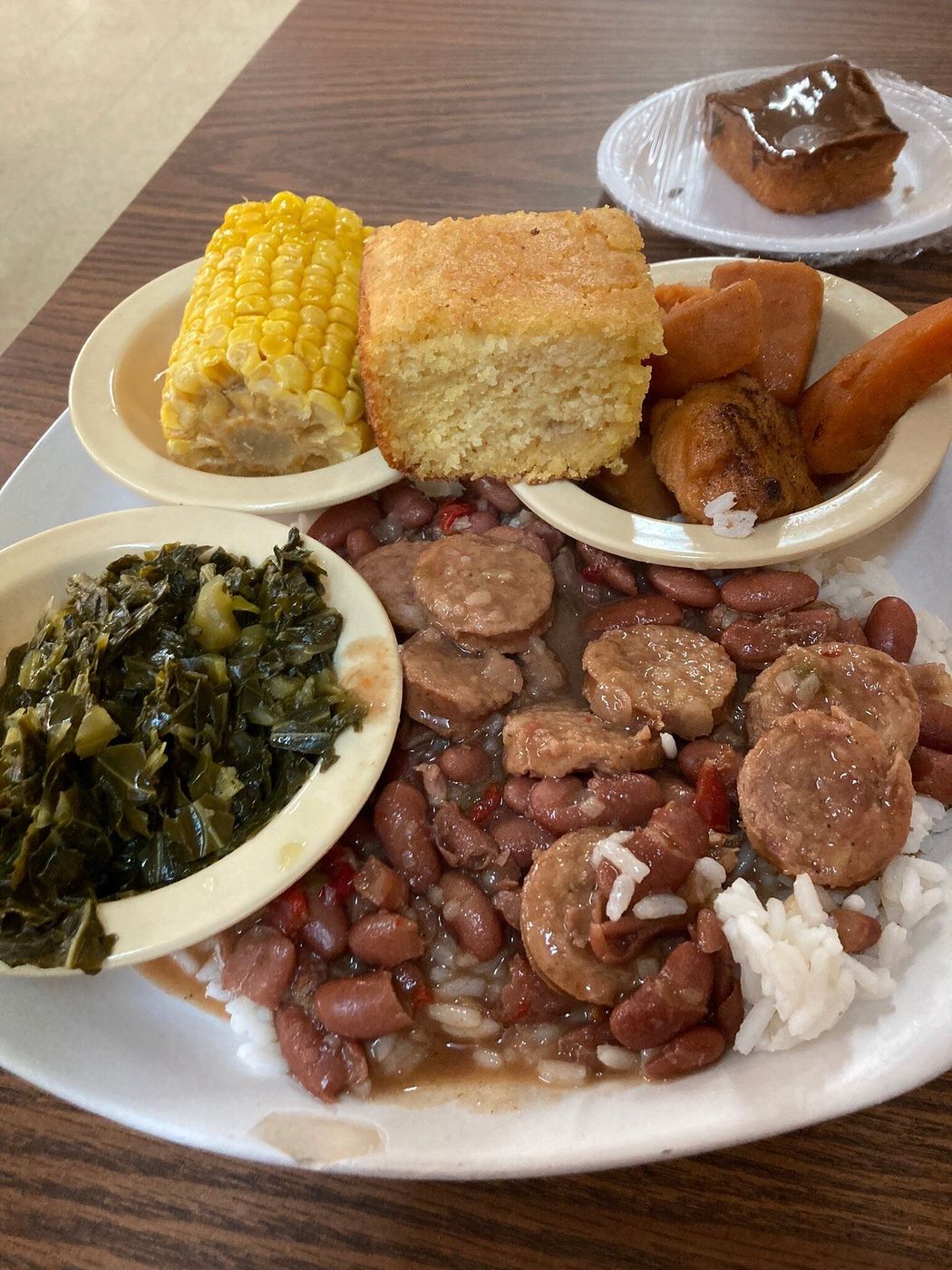 Plate lunch of red beans and rice with sausage, collard greens, corn on the cob, cornbread, yams and a slice of cake