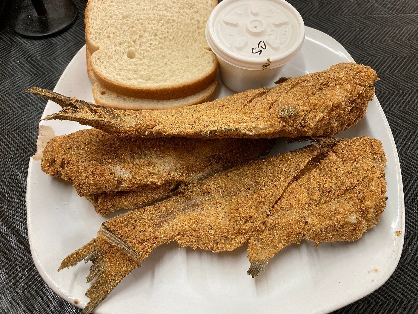 Three whole fried catfish on a white plate with sliced bread and a cup of slaw